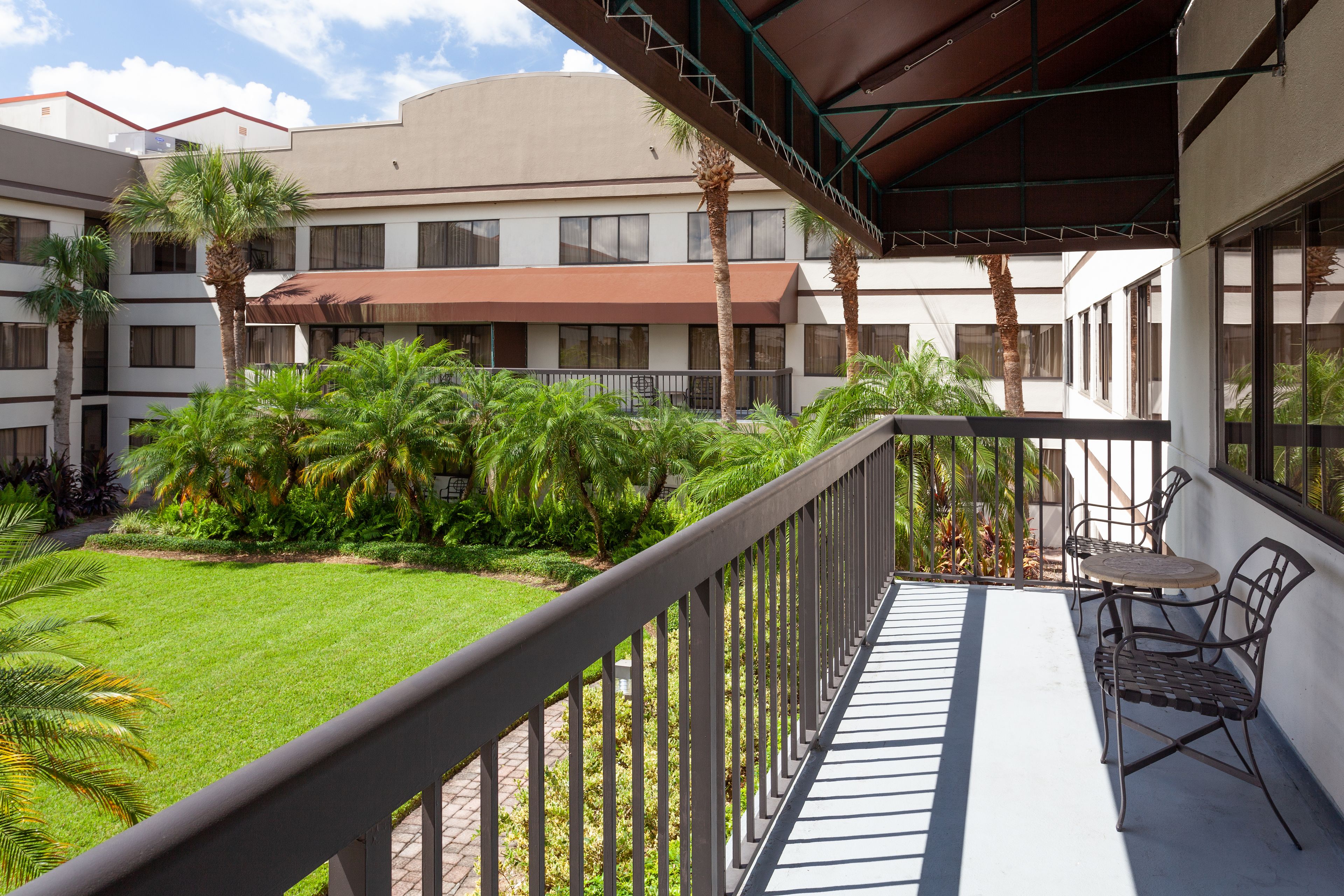 A balcony with a view of a well-maintained lawn and palm trees, a covered seating area with metal chairs, and a building with multiple windows and balconies in the background.