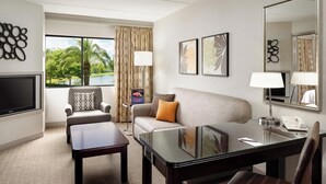 Une chambre d’hôtel équipée d’un canapé, d’un fauteuil, d’une table basse et d’un bureau avec une lampe. On y trouve également une télévision, un miroir et des œuvres d’art accrochées aux murs.