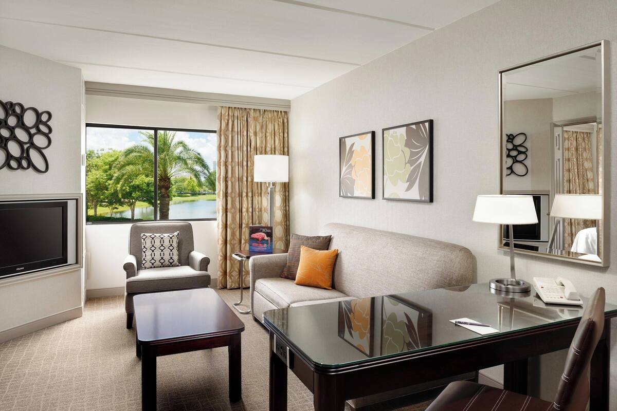 Habitación de hotel con sofá, sillón, mesa de centro y escritorio con lámpara. Hay un televisor, un espejo y obras de arte en las paredes.