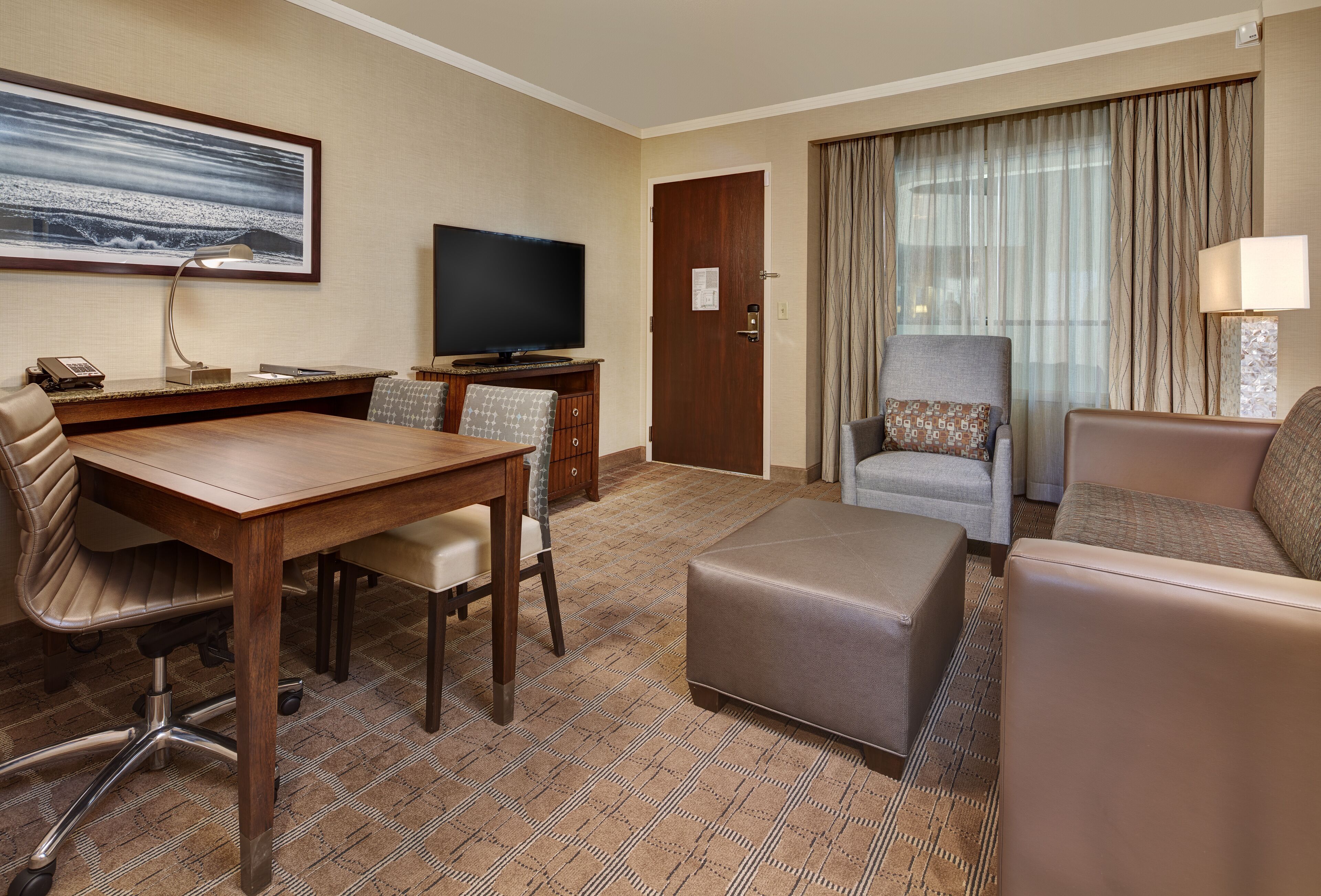 A hotel room with a wooden desk, a television, a sofa, and a large window with curtains.