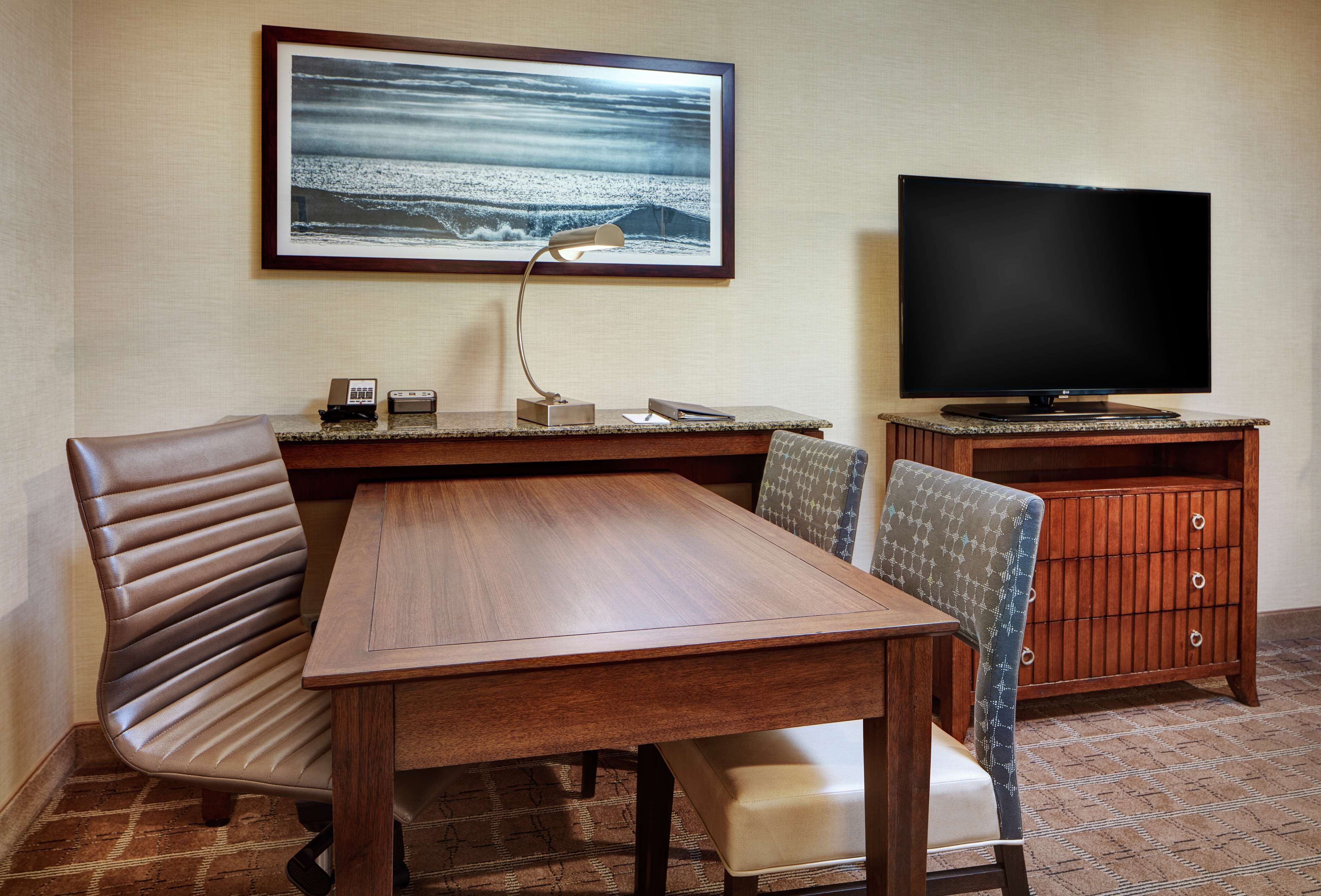 A hotel room with a wooden dining table, two chairs, a television, and a framed picture on the wall.