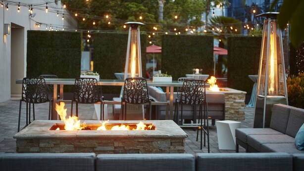 An outdoor patio with a stone fire pit, outdoor seating, string lights, and a tall, narrow heater.