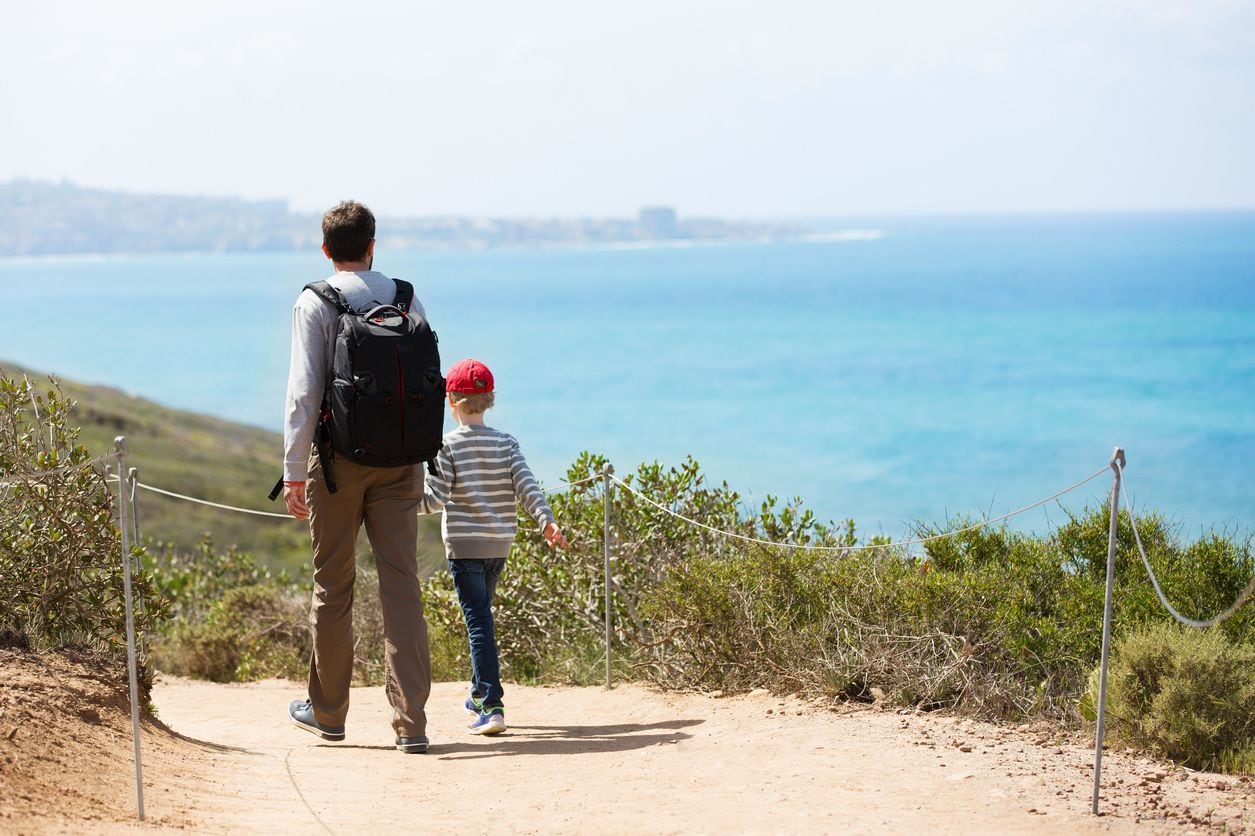 A man and a child walking on a dirt path with a scenic view of the ocean and distant city skyline.