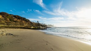 A beach with clear skies, a rocky cliff, and a lighthouse in the distance.
