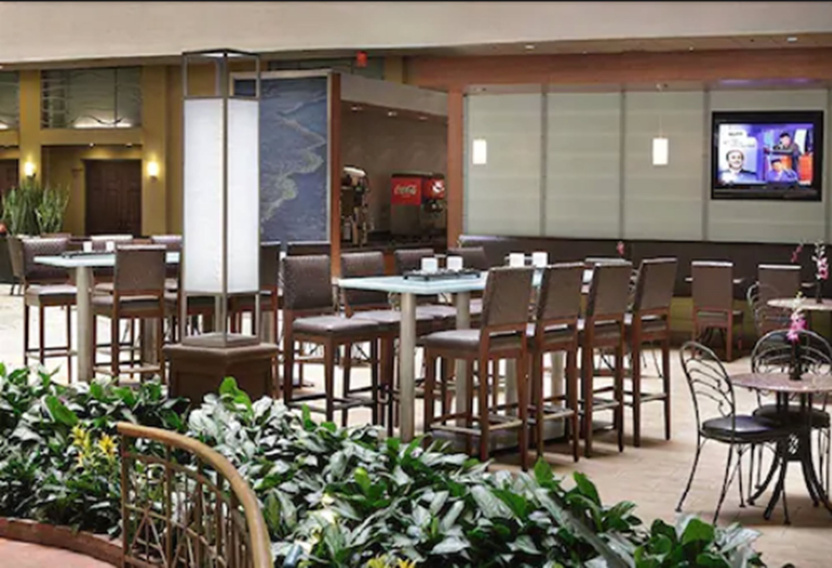 A restaurant with multiple tables and chairs, a television screen, and indoor plants.