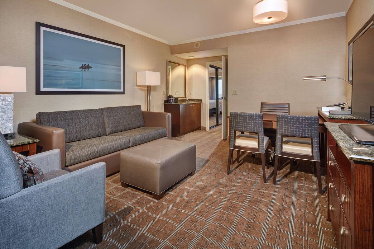 Ein Hotelzimmer mit Sofa, Sessel, Hocker, Essbereich mit Stühlen, einer kleinen Küchenzeile und einem großen, gerahmten Bild an der Wand.