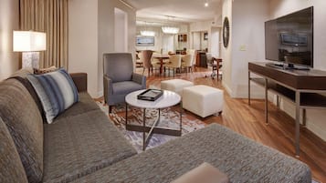 Una habitación de hotel moderna con un sofá gris, una pequeña mesa redonda, un televisor y un área de comedor al fondo.