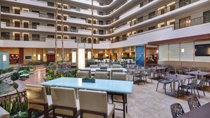 A multi-level hotel lobby with dining areas, a reception desk, and a large TV screen.