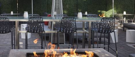 An outdoor dining area with a fire feature, tables, chairs, and red umbrellas.