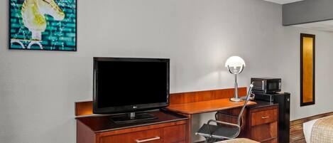 A hotel room with a flat-screen TV, a desk with a lamp, a microwave, and a bed with patterned bedding.