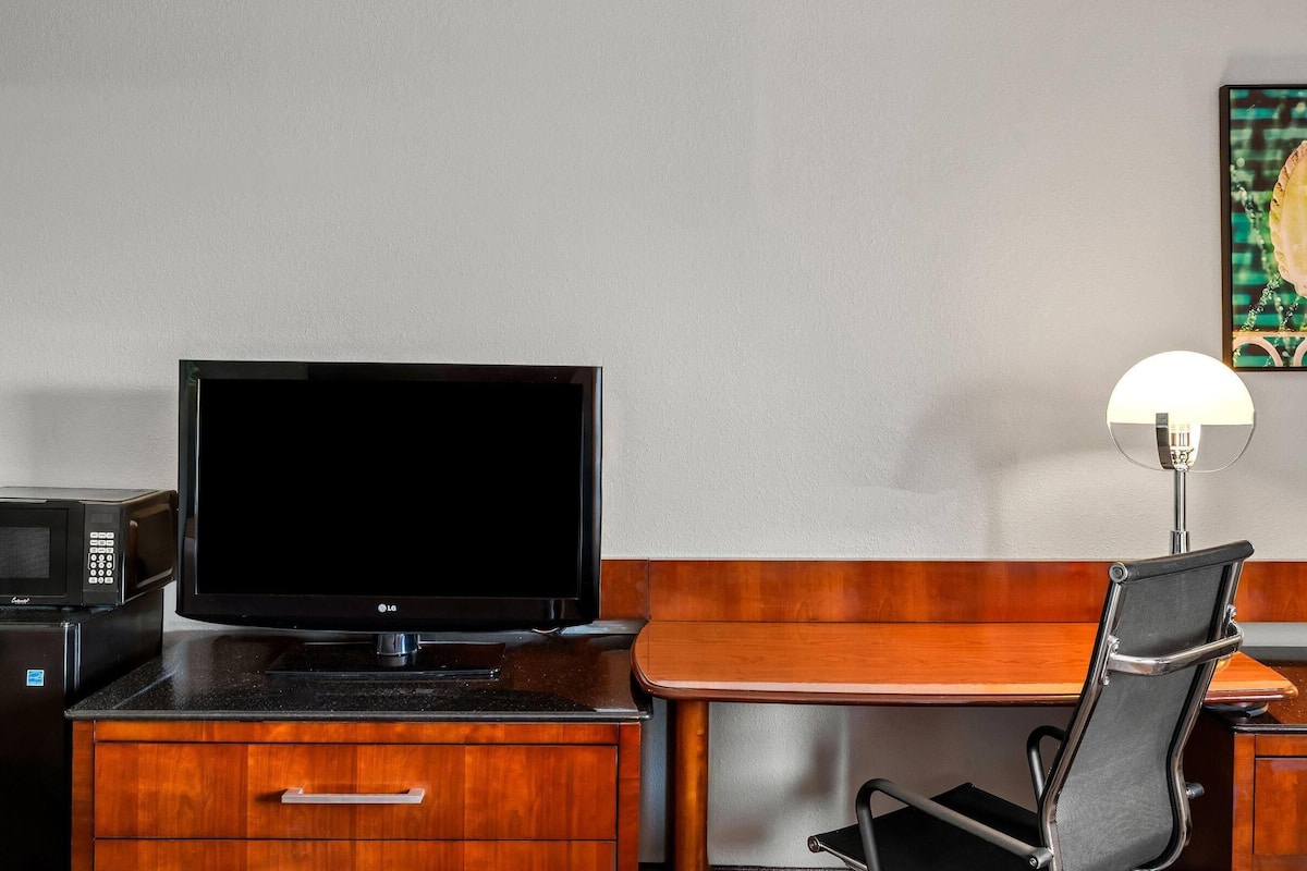 A hotel room with a wooden desk, a black office chair, a flat-screen TV, a microwave, and a lamp.