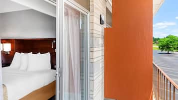 A hotel room with a bed, a sliding glass door, and a view of a parking lot and greenery.