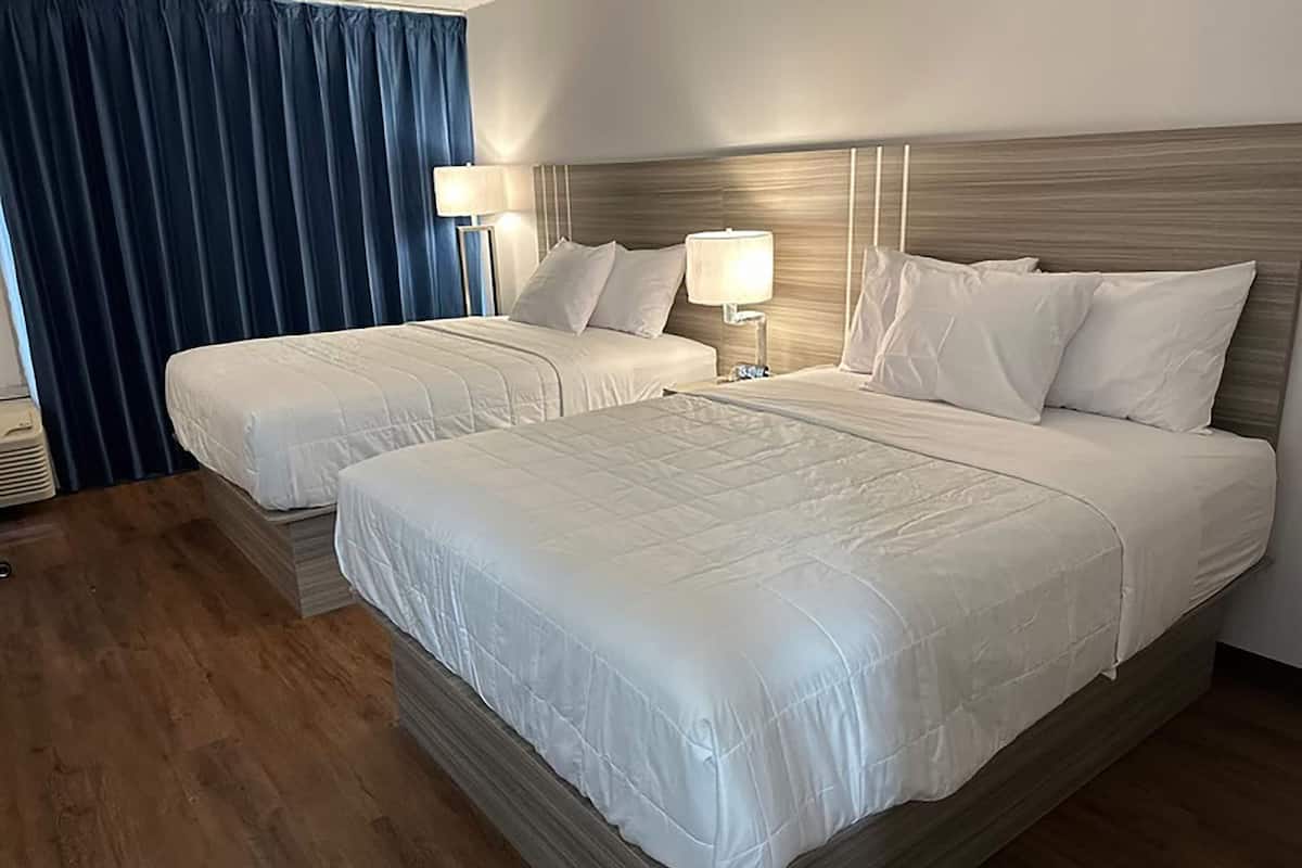 A hotel room with two beds, a wooden headboard, a nightstand with a lamp, and a blue curtain.