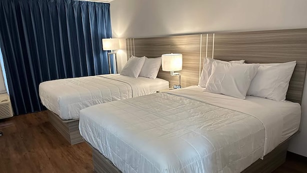 A hotel room with two beds, each with white bedding and headboards. There are bedside lamps and a large window with blue curtains. - Baymont by Wyndham Milesburg (Howard)