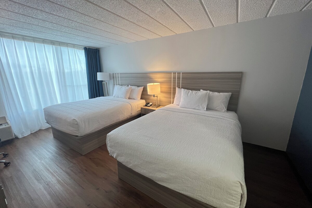 Habitación de hotel con dos camas, una mesita de noche con lámpara y una ventana con cortinas.