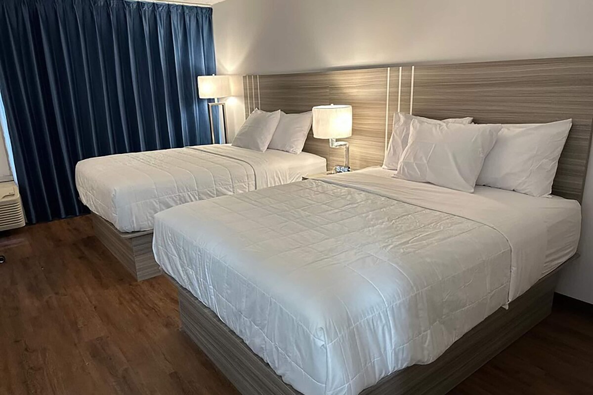 A hotel room with two beds, a wooden headboard, a nightstand with a lamp, and a blue curtain.