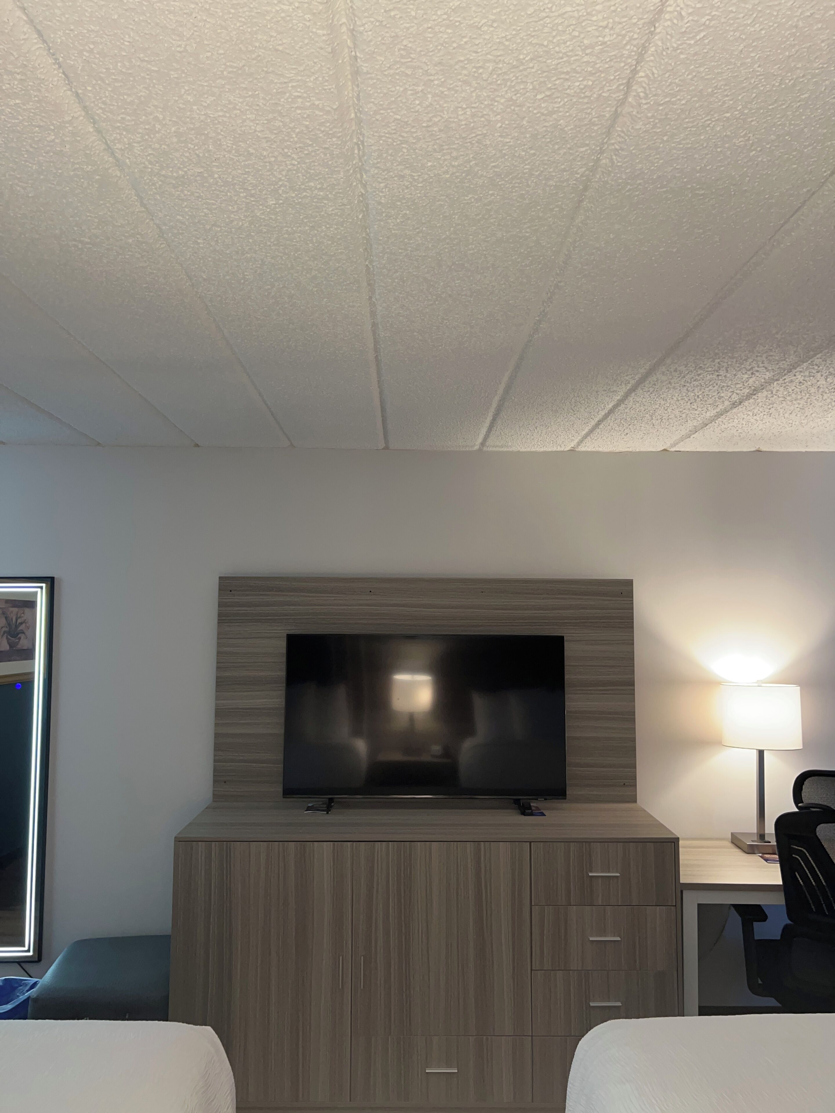A hotel room with a flat-screen TV mounted on a wooden cabinet, a desk with a lamp, and two beds.