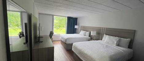 A hotel room with two beds, a desk, a chair, and a large window with a view of greenery.