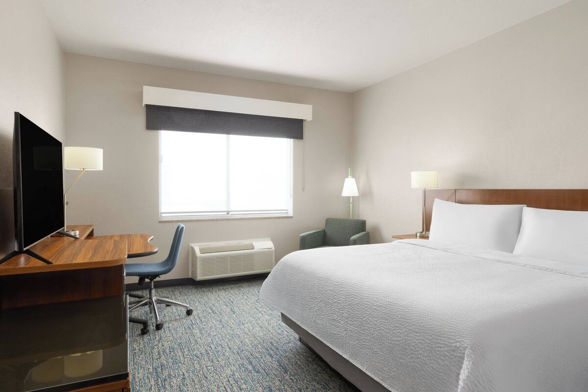 A hotel room with a bed, a desk with a TV, a chair, a lamp, and a window with blinds.
