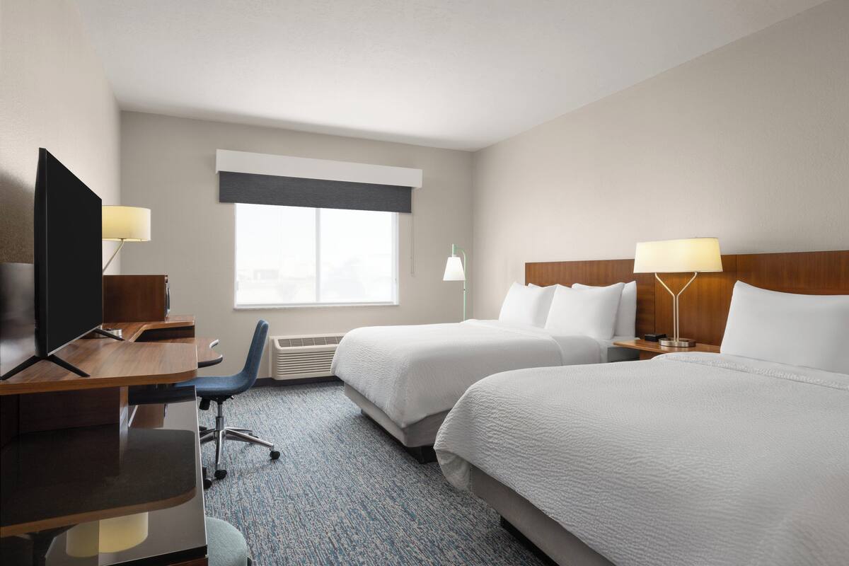 A hotel room with two beds, a desk, a chair, a TV, and two lamps.