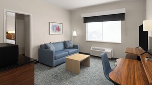 A hotel room with a blue sofa, a wooden coffee table, a TV, a desk, and a window with a black blind.