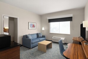 A hotel room with a blue sofa, a wooden coffee table, a TV, a desk, and a window with a black blind.