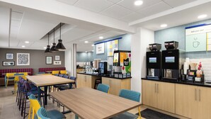 Een moderne eethoek met een lange houten tafel en stoelen, een keukenbar met koffieapparaten, en een zithoek met een rode, beklede bank.