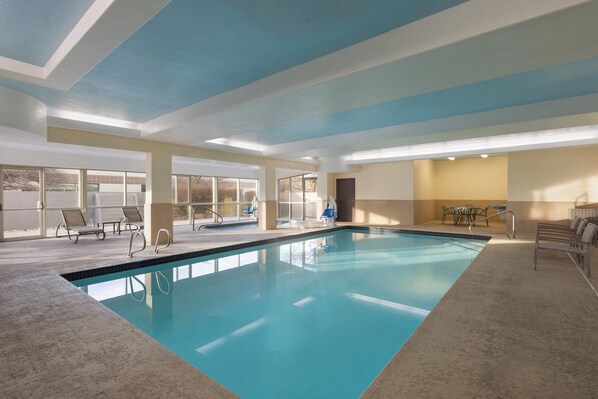 An indoor swimming pool with clear water, surrounded by lounge chairs and a view of the outside.