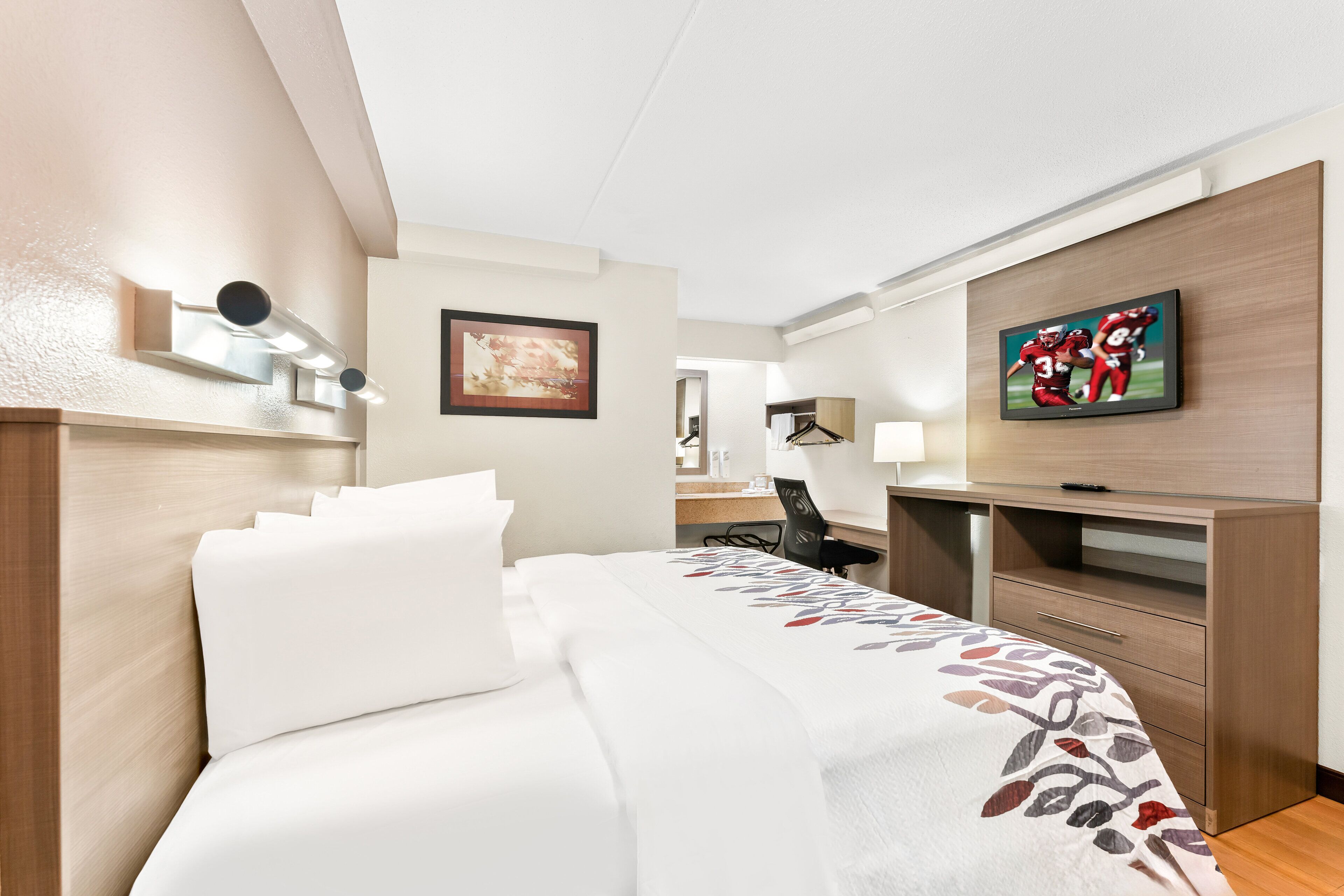 A hotel room with a bed, a desk, a chair, a TV, and a framed picture on the wall.