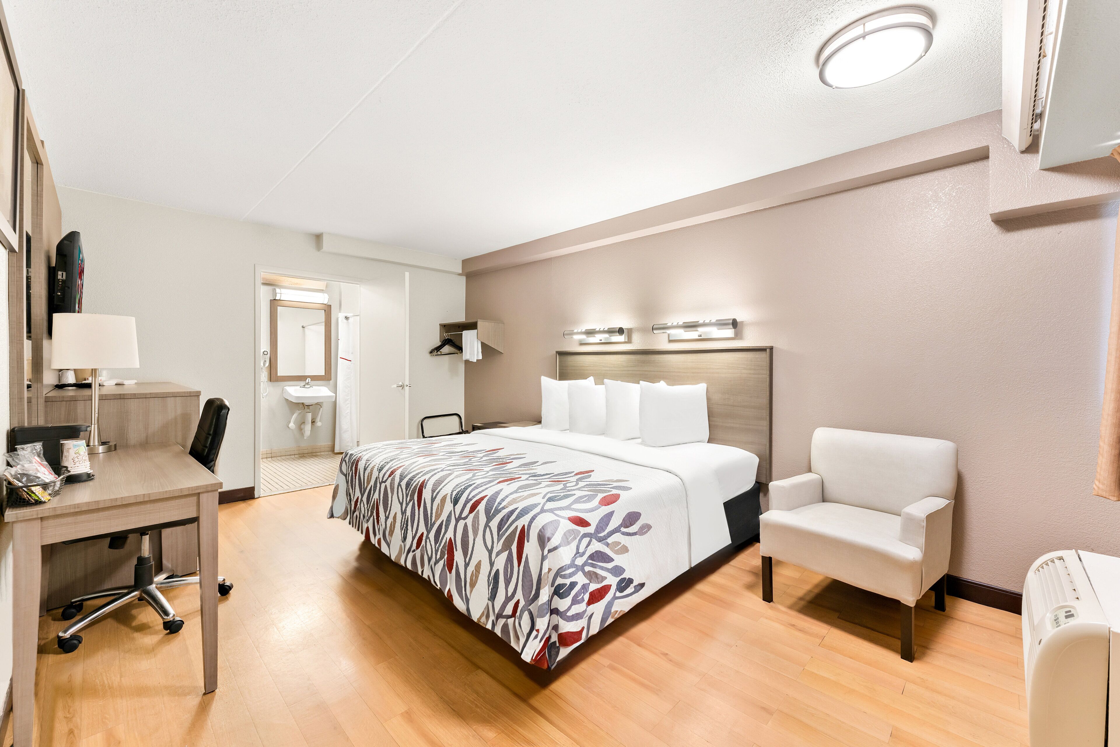 A hotel room with a bed, a desk, a chair, a bedside table, a lamp, a mirror, and a wall-mounted heater.