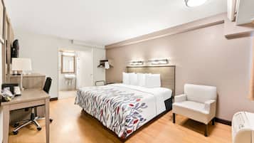 A hotel room with a bed, a desk, a chair, a bedside table, a lamp, a mirror, and a wall-mounted heater.