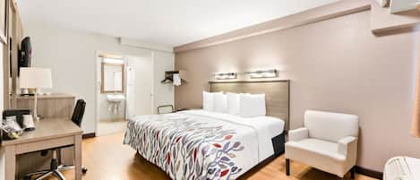 A hotel room with a bed, a desk, a chair, a bedside table, a lamp, a mirror, and a wall-mounted heater.
