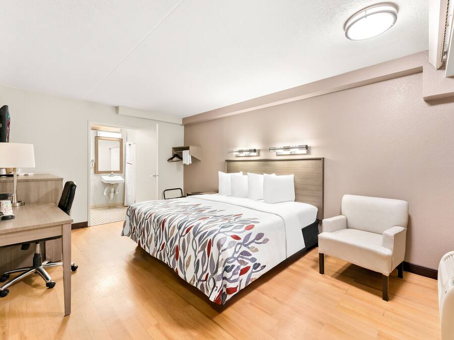 Quarto de hotel com cama, escrivaninha, cadeira, mesa de cabeceira, abajur, espelho e aquecedor fixado na parede.