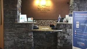 Ein Empfangsschalter mit einer Wand aus Stein im Hintergrund, ein Schild für 'Quality Inn & Suites' und ein Werbeständer für das 'Choice Rewards Program'.