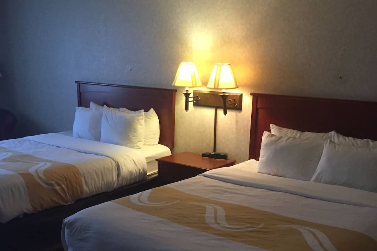 A hotel room with two beds, each with white linens and a wooden headboard. There is a nightstand between the beds with two lamps.
