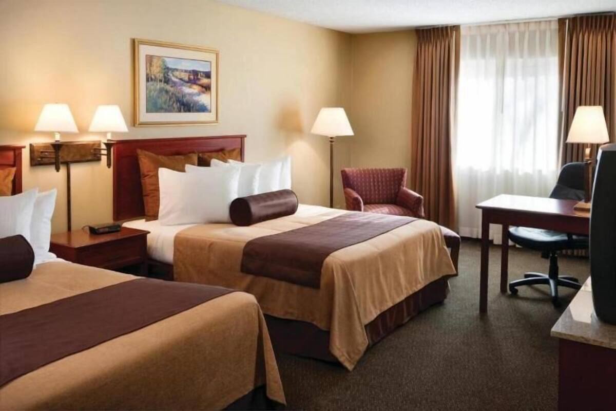 A hotel room with two beds, a desk, a chair, lamps, and a painting on the wall.