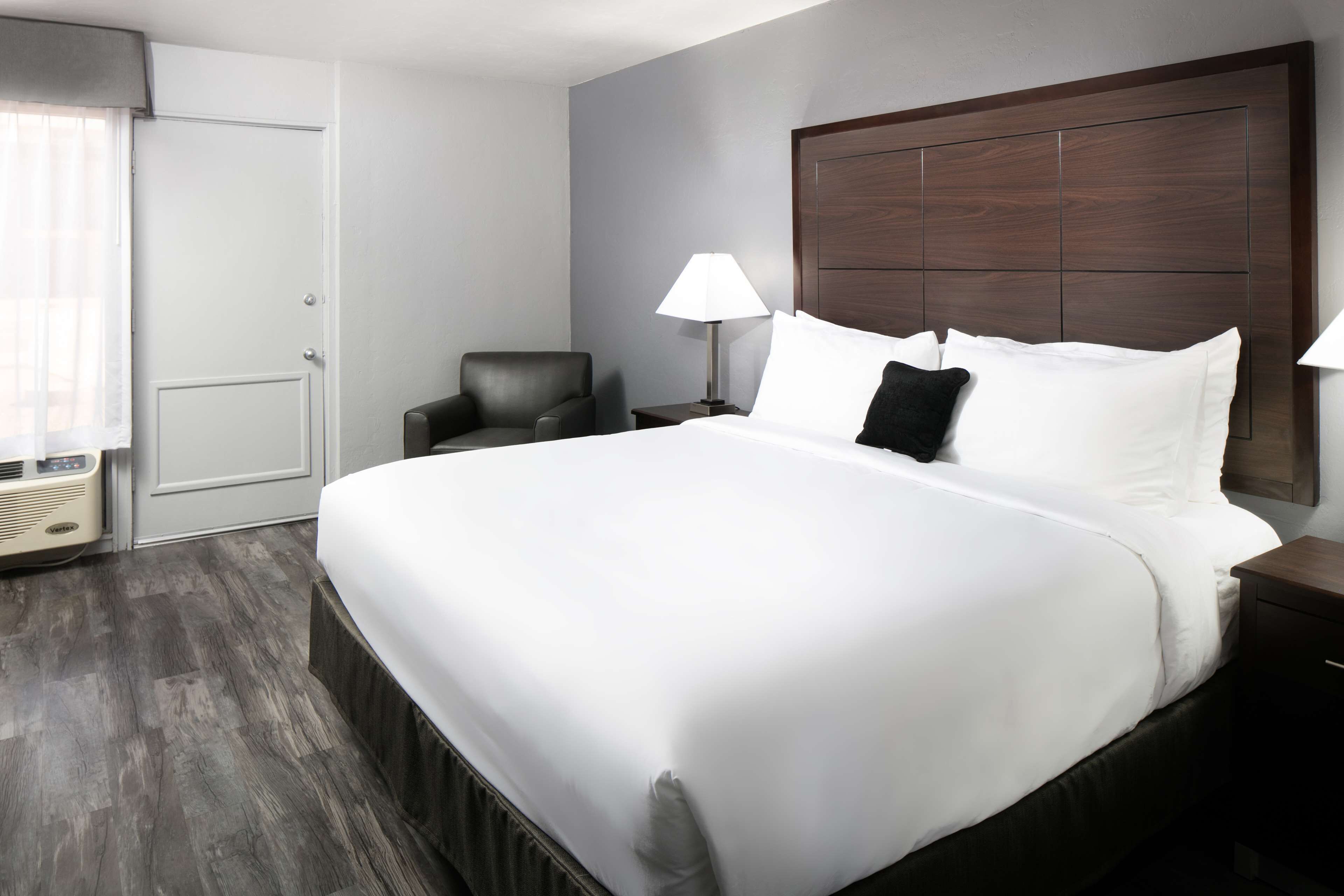 Ein Hotelzimmer mit einem großen Bett, einem Sessel, einem Nachttisch, einer Lampe und einem Fenster mit Jalousien.