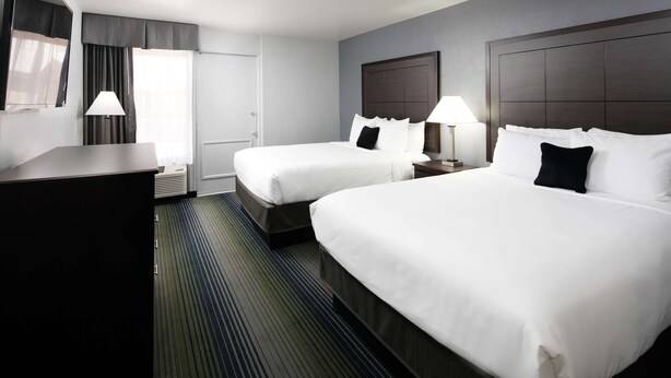 Quarto de hotel com duas camas, uma escrivaninha, uma cadeira, um abajur e uma janela com cortinas.