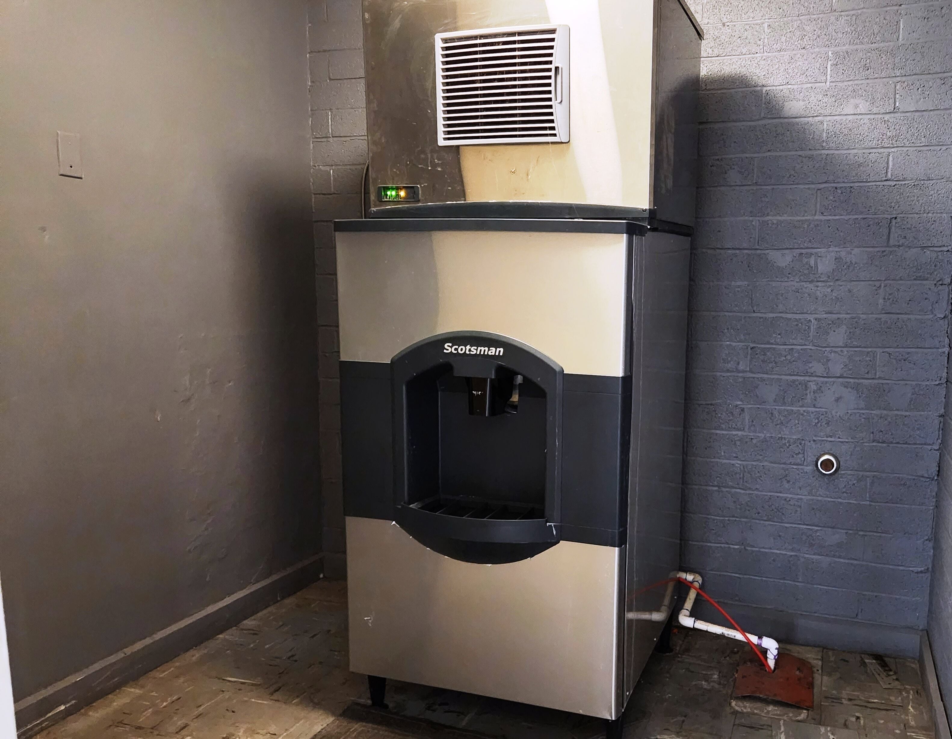 A compact, stainless steel ice maker with a digital display and a water dispenser, positioned against a grey brick wall in a corner.