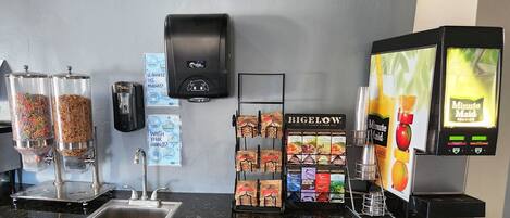 A self-service station with dispensers for cereal and granola, a hand sanitizer dispenser, a soap dispenser, a handwashing instruction sign, a Bigelow tea display, and a Minute Maid juice dispenser.