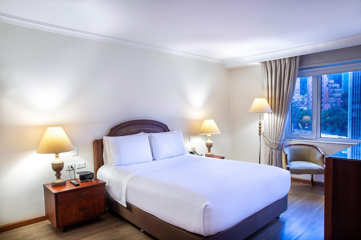 Une chambre d’hôtel avec un grand lit, deux tables de chevet avec des lampes, une chaise et une fenêtre donnant sur la ville.