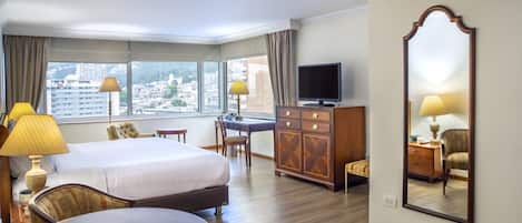 Habitación de hotel con cama, escritorio, silla, cómoda, televisor y ventanas amplias que dan a vistas de la ciudad.