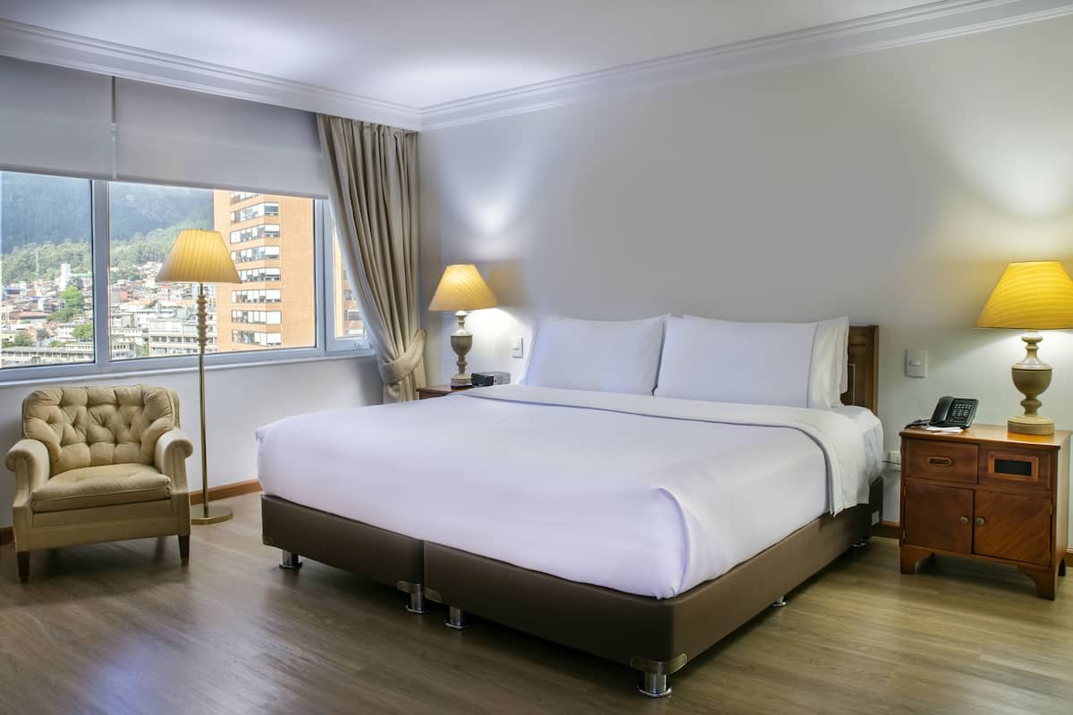 Une chambre d’hôtel avec un grand lit, une chaise, une table de chevet avec une lampe et une vue sur le paysage urbain par la fenêtre.