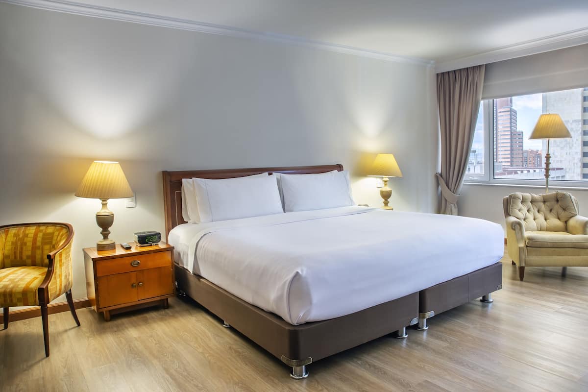 Une chambre d’hôtel avec un grand lit, deux tables de chevet avec des lampes, une chaise et une vue sur la ville par la fenêtre.