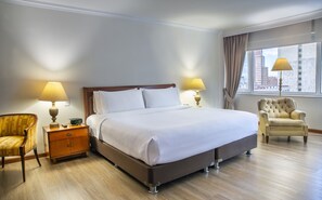 Habitación de hotel con una cama grande, dos mesitas de noche con lámparas, una silla y vistas a la ciudad por la ventana.