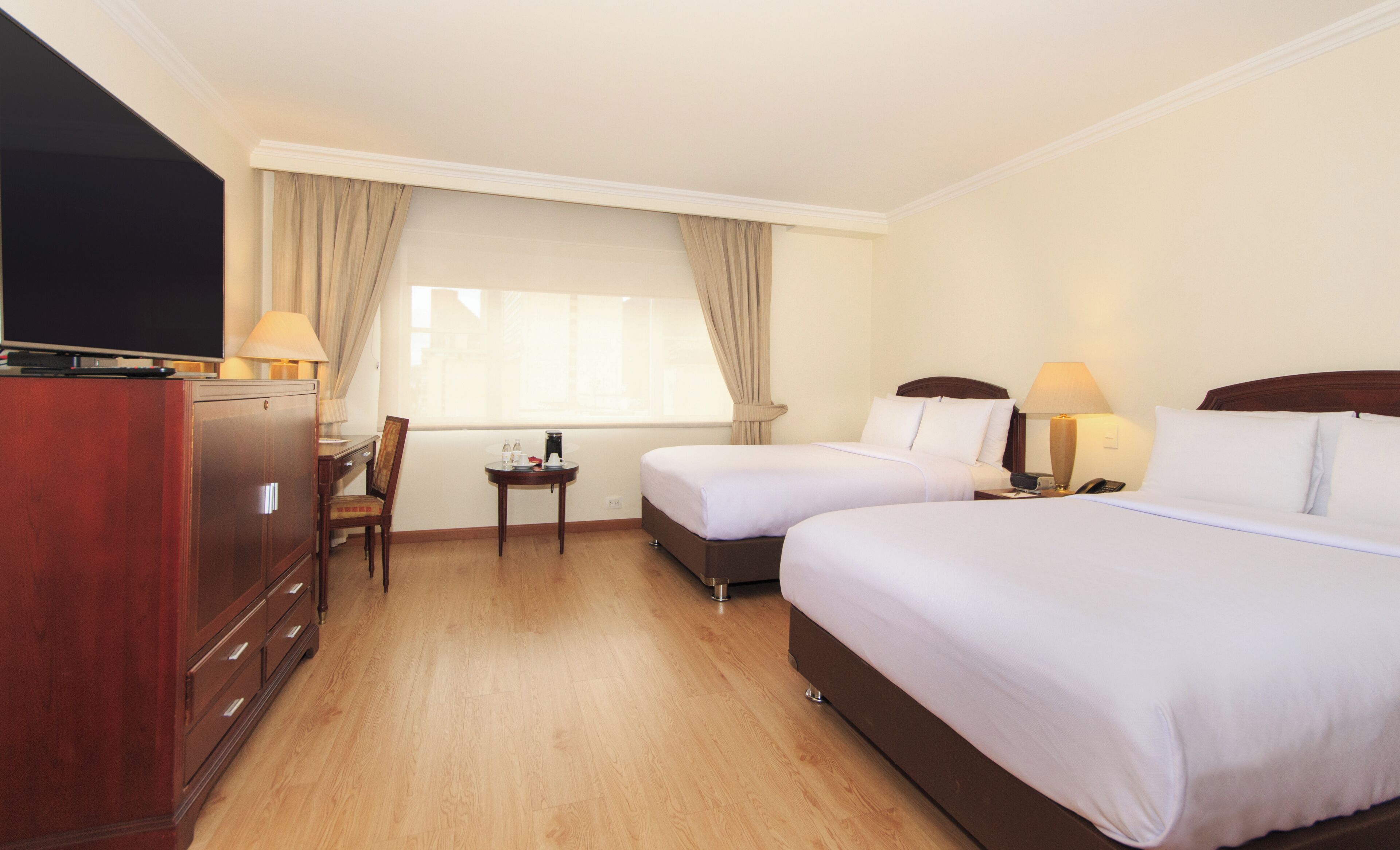 A hotel room with two beds, a TV, a desk with a chair, a small table, and a window with curtains.