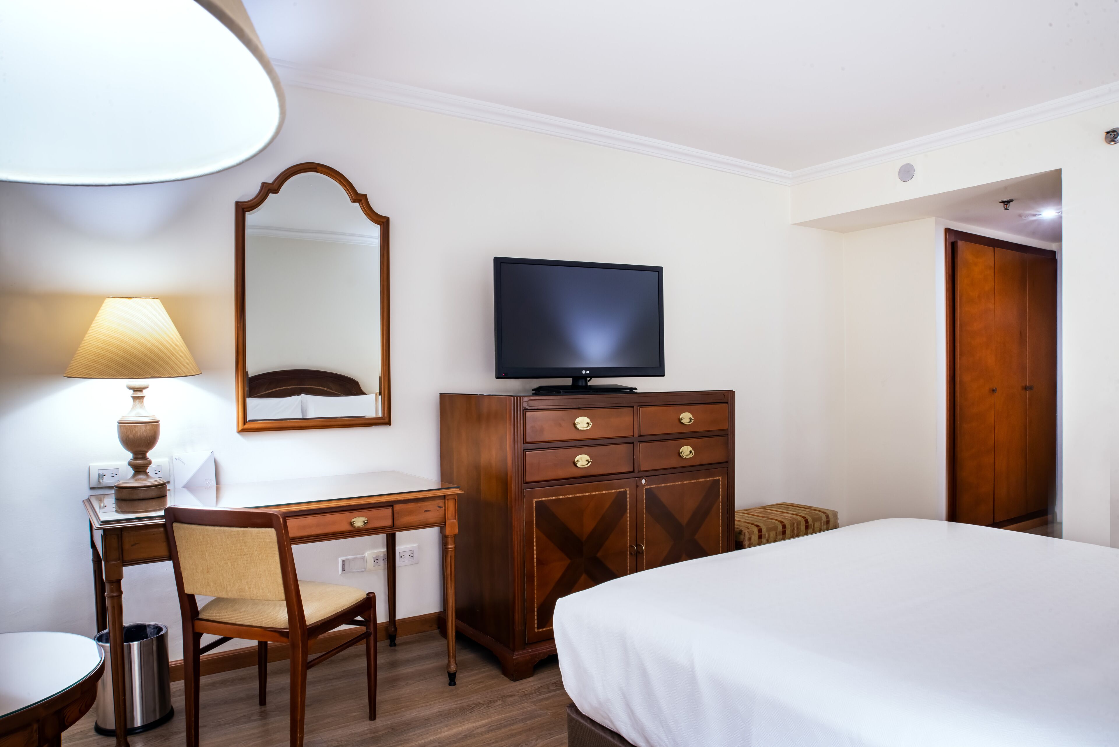 Habitación de hotel con cama, escritorio y silla, televisor, cómoda, lámpara y espejo.