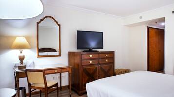Habitación de hotel con cama, escritorio y silla, televisor, cómoda, lámpara y espejo.