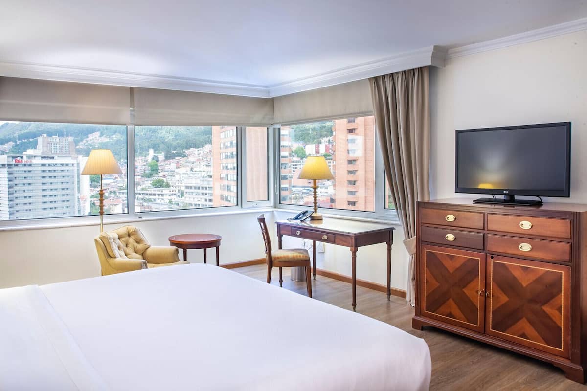 Une chambre d’hôtel équipée d’un lit, d’un bureau, d’une chaise, d’une télévision et de grandes fenêtres avec des rideaux.