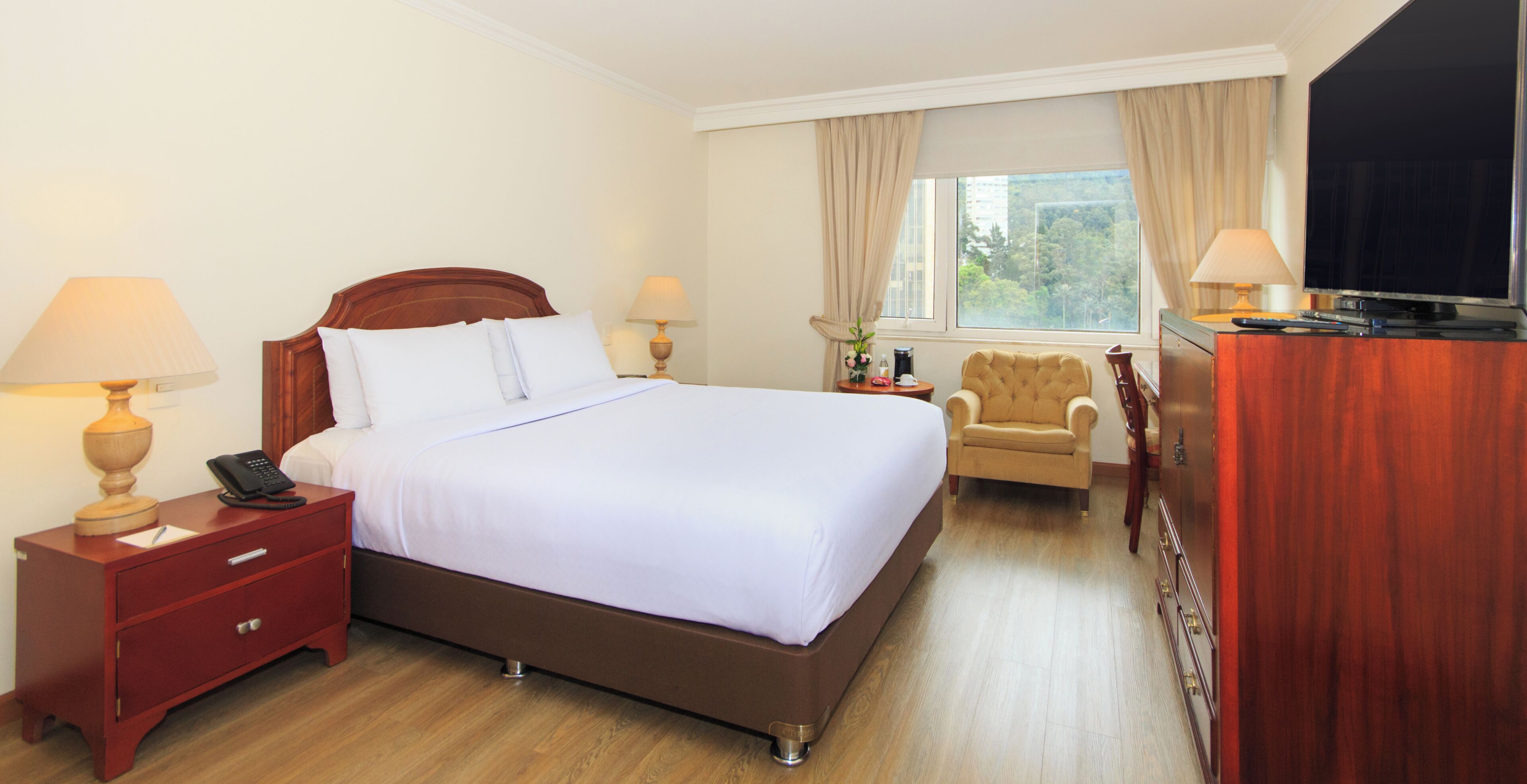 A hotel room with a large bed, bedside tables with lamps, a chair, a desk with a TV, and a window with curtains.